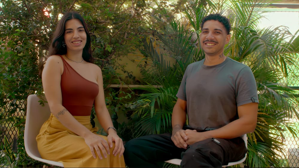 Yasmim e Igor, a dupla da Troça, sentados entre plantas